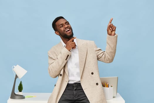 Happy energetic man in business casual clothes standing in bright contemporary office, gesturing with finger upward, cheerful mood, blue backdrop, workspace with computer, stationery, and plant, work life style concept.