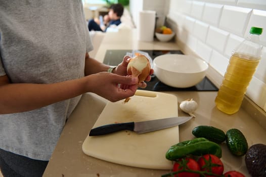 A person peels an onion in a contemporary kitchen with fresh ingredients laid out, including tomatoes and cucumbers. A detail view emphasizing culinary preparation and healthy eating.