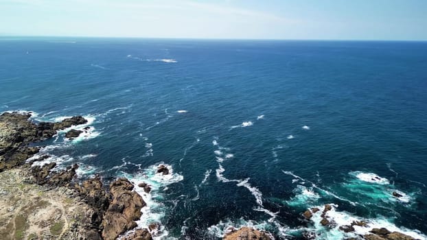 Breathtaking aerial footage of the ocean waves crashing against rocky shores under a clear blue sky.