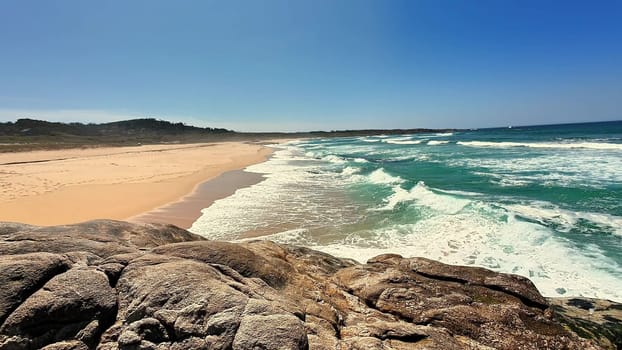 A breathtaking view of a beautiful sandy beach with waves widely crashing against rocks beneath clear blue skies