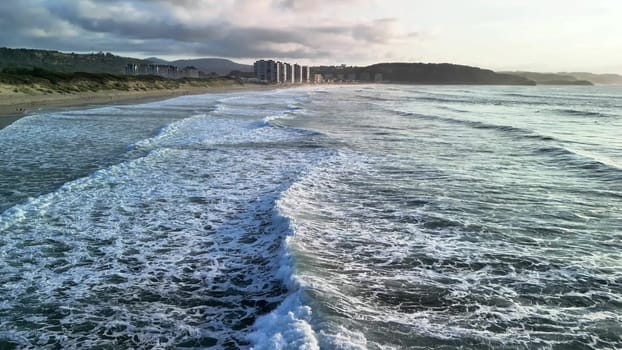 Beautiful waves are crashing gently on a serene beach while distant buildings stand under a cloudy sky