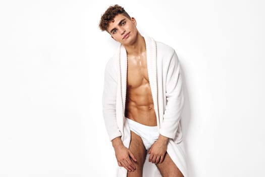 Young muscular man in a white bathrobe exuding confidence and relaxation. The pure white background emphasizes the theme of wellness and personal grooming.