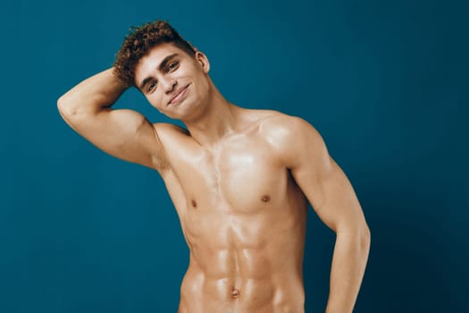 Confident young man with toned abs and curly hair, smiling as he poses against a solid blue background. Emphasizing a fitness and wellness lifestyle.