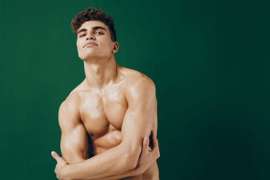 Athletic man displaying confidence with crossed arms, glistening skin and a strong build. Green backdrop emphasizes fitness and personal strength.