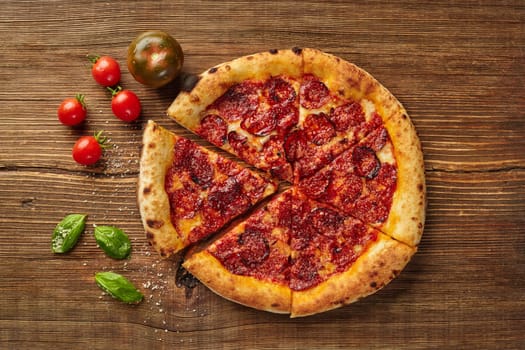 Sliced pepperoni pizza sits on a wooden table, surrounded by fresh tomatoes and basil leaves