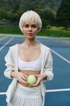 Confident young woman with short blonde hair in sportswear holds a tennis ball, standing on a sunny outdoor tennis court surrounded by greenery