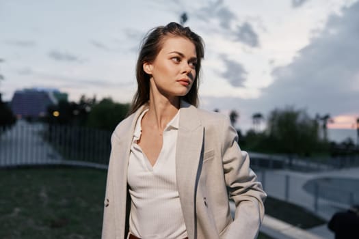 Thoughtful woman wearing beige business suit and white shirt stands outdoors in urban setting with soft natural sunset lighting and cloudy blue sky, evoking business confidence and professionalism.