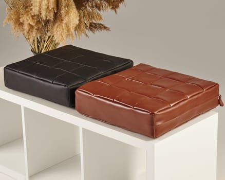 Two square pouf cushions, one brown and one black, made of patterned leather, sit on a white, minimalist shelf