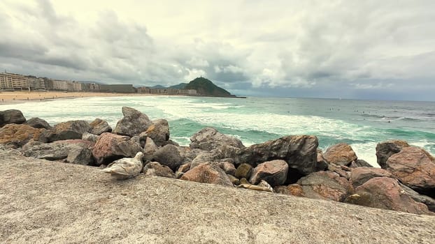 Experience a tranquil beach scene that features rugged rocky shores and dynamic waves cascading under overcast skies