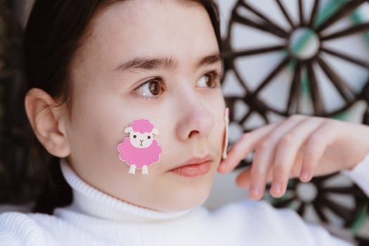 Portrait of a beautiful Caucasian brunette girl with brown eyes and a pink lamb sticker on her cheek, looking away while sitting in a hanging swing in the garden on a spring day, side view close-up. Concept children creativity, happy Easter, leisure child.