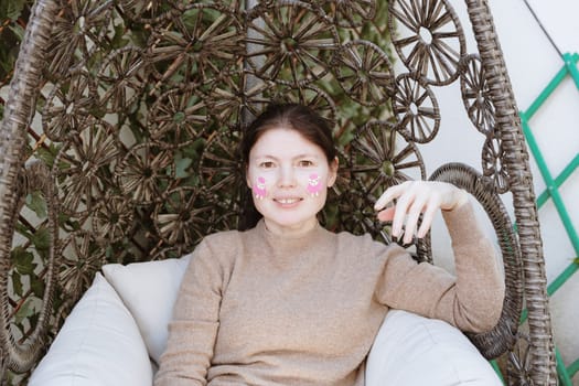 Portrait of one beautiful Caucasian young brunette woman with brown eyes and two pink lamb stickers on her cheeks, who is fooling around and looking at the camera while sitting in a hanging swing in the garden on a spring day, side view close-up. Concept of a woman, creativity, happy Easter, leisure, people.