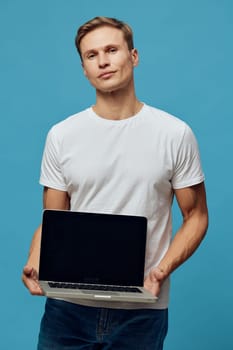 Friendly young man with casual white t-shirt holding a laptop computer isolated on a plain blue background, emphasizing modern technology and professionalism. Concept: digital skills and online work.