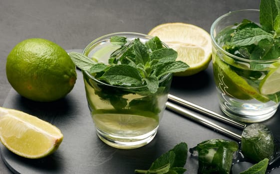 Glass of mojito cocktail with fresh mint leaves, lime slices, and ice cubes on a dark stone background. Refreshing summer drink with stainless steel straws