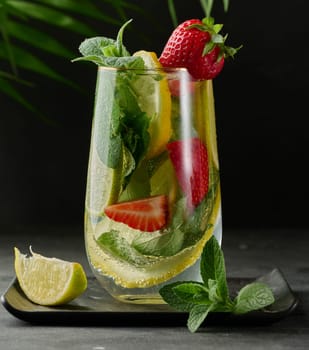 A refreshing drink in a glass with lemon slices, strawberries, fresh mint leaves, and sparkling water, served with lime and mint on a black plate.