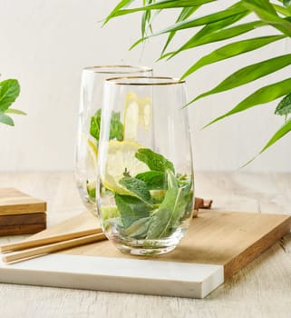 Preparing a refreshing drink. Two glasses filled with fresh lemon slices and mint leaves on a wooden board with a green plant background