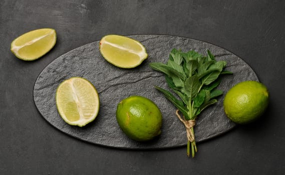 Whole and cut limes with fresh mint leaves arranged on a dark textured surface. Composition perfect for food, drink, or summer themes