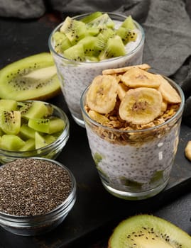 Two glasses of chia seed pudding, one topped with diced kiwi and the other with granola and banana chips, are on a dark surface with ingredients