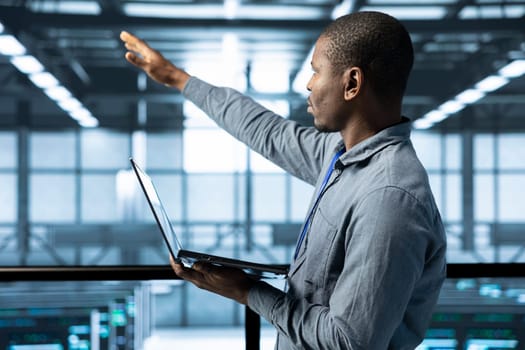 Technician holding laptop, looking around data center, managing server virtualization, finetuning network protocols, verifying high availability configurations, enforcing advanced security measures