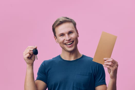 smiling young caucasian man wearing blue t-shirt hold car key and brown envelope look camera on pink background with happy emotion. This digital nomad concept symbolizes freedom, remote work and mobility lifestyle.
