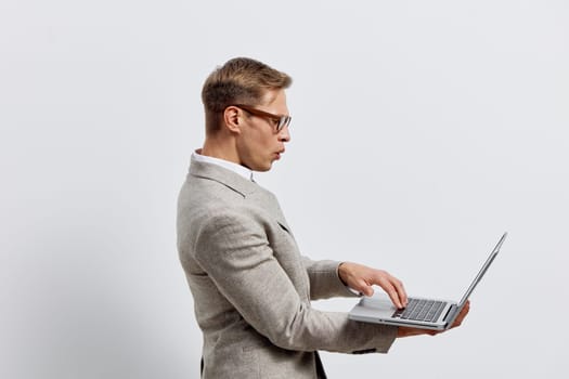 surprised man holding laptop wearing glasses and gray blazer on plain white background. young caucasian man with casual hairstyle using laptop for work in digital nomad lifestyle concept. innovation, remote work, connectivity.