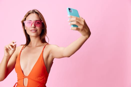 Vibrant woman in orange swimsuit capturing a selfie on her cellphone against a soft pink backdrop