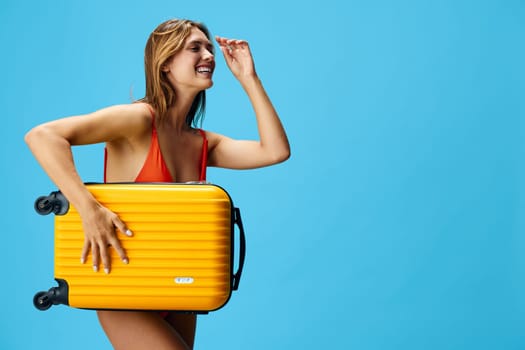 Vibrant summer getaway young woman in colorful swimsuit poses with yellow suitcase against serene blue backdrop