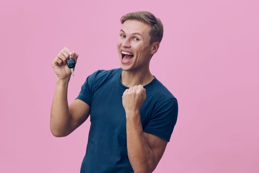 Cheerful man in casual blue t-shirt holding car key, showing excitement and happiness. Vibrant pink background emphasizes positive mood, lifestyle, success, achievement, celebration, joy.