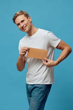 Cheerful young man with blond hair in casual clothing holding a blank cardboard sign and smiling at the camera against a vivid blue background. Concept: positive communication, youth lifestyle, studio portrait, vibrant colors, happiness.