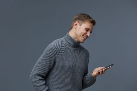 Happy man with short hair in cozy gray knit sweater looking at smartphone with smile against simple gray backdrop. Concept: enjoyment of digital communication, relaxed lifestyle, modern technology.