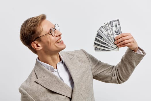 Happy professional man with glasses displaying cash of hundred dollar bills, wearing beige blazer and white shirt, looking to the side with a confident expression, isolated on light background. Concept: success and wealth.