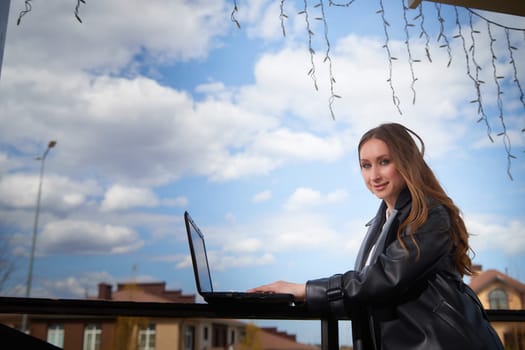 A young woman sits at a table using a laptop while enjoying a beautiful day outdoors under a partly cloudy sky.