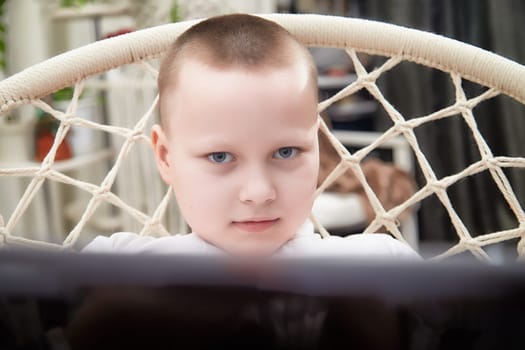 A young boy intently stares at a computer screen while comfortably seated in a macrame chair at home in the afternoon.