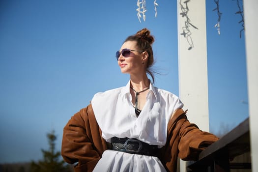 A fashionable woman stands confidently outdoors, basking in the sun while wearing stylish sunglasses and a unique outfit.