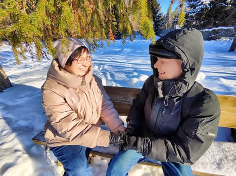 Two smiling individuals sit on a bench, holding hands in a snowy park. Sunshine filters through evergreen trees, creating a serene winter atmosphere. Joy fills their expressions.