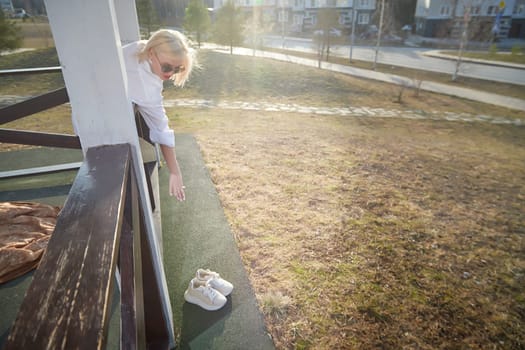 A person leans over a balcony with sunglasses, extending their arm toward a pair of shoes placed on the ground below in bright sunlight.
