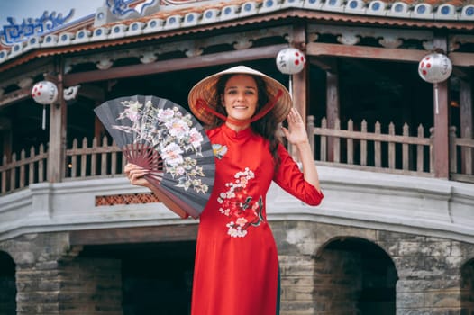 female traveler tourist in a Vietnamese hat with a fan at the ancient Japanese covered bridge in old town in Hoi An in Vietnam in Asia