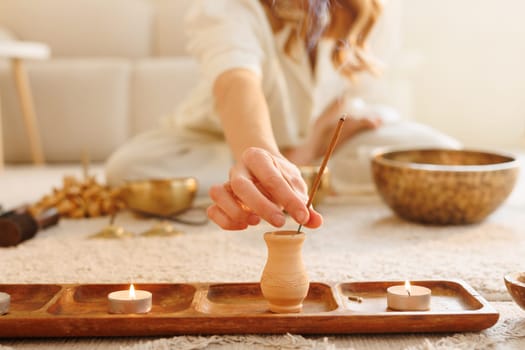 Woman lighting incense stick in cozy space with candles and Tibetan bowls for meditation and energy cleansing.