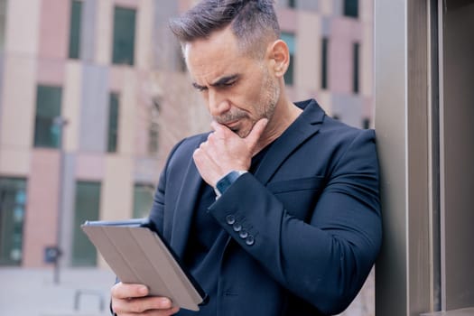 Businessman leaning on wall, using a tablet with a thoughtful expression. He wears a black suit and smartwatch in an urban setting. Concept of focus, planning, and modern digital lifestyle.
