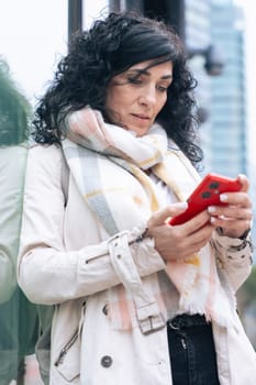 Happy caucasian woman standing outdoors in urban setting, wearing coat and scarf, using her red smartphone. She is smiling and relaxed, enjoying a casual moment on a cold day in the city. Vertical