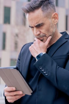 Businessman leaning on wall, using a tablet with a thoughtful expression. He wears a black suit and smartwatch in an urban setting. Concept of focus, planning, and modern digital lifestyle. Vertical