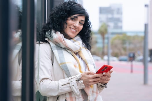 Happy caucasian woman standing outdoors in urban setting,wearing coat and scarf,using her red smartphone.She is smiling and relaxed,enjoying a casual moment on a cold day in the city looking at camera