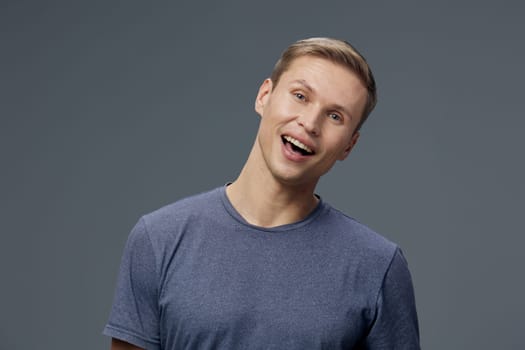 cheerful smiling man with short blond hair wearing gray casual t-shirt tilting head and looking at camera with upbeat facial expression against plain gray background digital detox concept promoting mental health and gadget balance