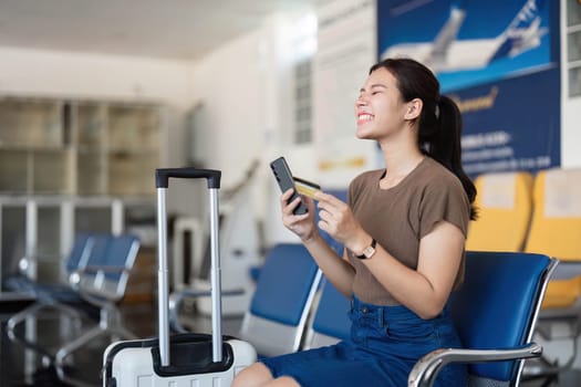 A happy young woman seated at the airport, excitedly using her phone and suitcase, capturing the spirit of travel anticipation.