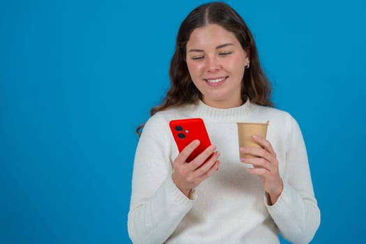 Portrait of a smiling young woman in white sweater holding a red smartphone and a coffee cup against a blue background, representing communication, relaxation, and casual modern lifestyle.Copy space