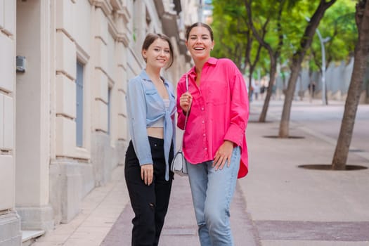 Caucasian women smiling closely while taking a selfie together on a city street. Outdoor setting with trees and buildings, capturing a joyful and candid moment of friendship and connection