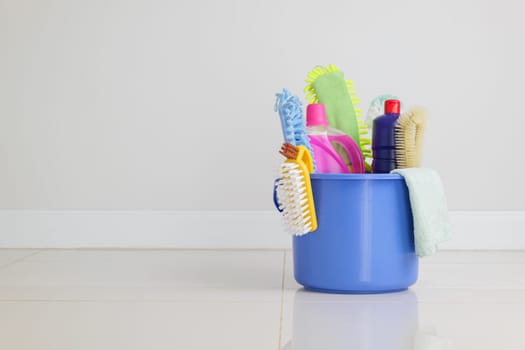 Cleaning routine essentials. blue plastic bucket filled with detergent bottles, brushes, and other cleaning supplies sits on the floor in the living room, prepared for housekeeping and daily housework