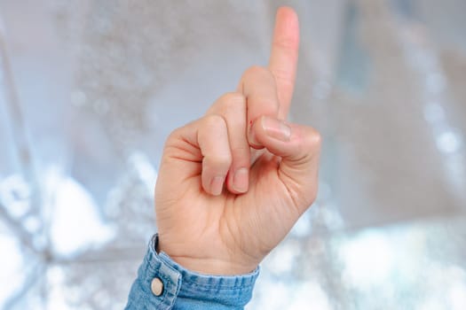 close-up image of a human hand wearing a denim shirt, raising a single finger upward in a pointing gesture, with a soft reflective background, symbolizing direction, focus, or instruction.