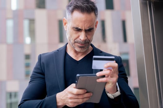 Confident caucasian man with headphones serious while using tablet and holding a credit card outdoors. Concept of online shopping, digital payment, modern lifestyle, and financial technology.