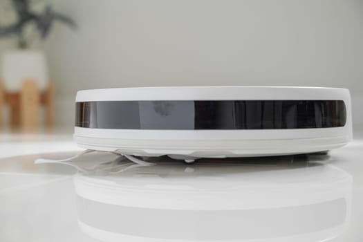 White automatic floor cleaner with side brush on tile flooring, featuring front black sensor strip. Robot vacuum cleaner efficiently cleaning floor, illustrating smart home technology for dust removal