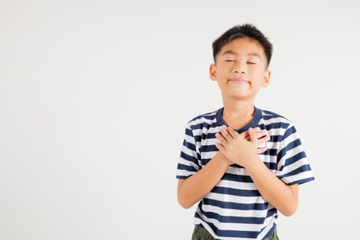 Happy young kid boy in casual clothes holding hands at chest close to heart meditation and smiling in studio short isolated on white background, primary child believe faith and love gratitude